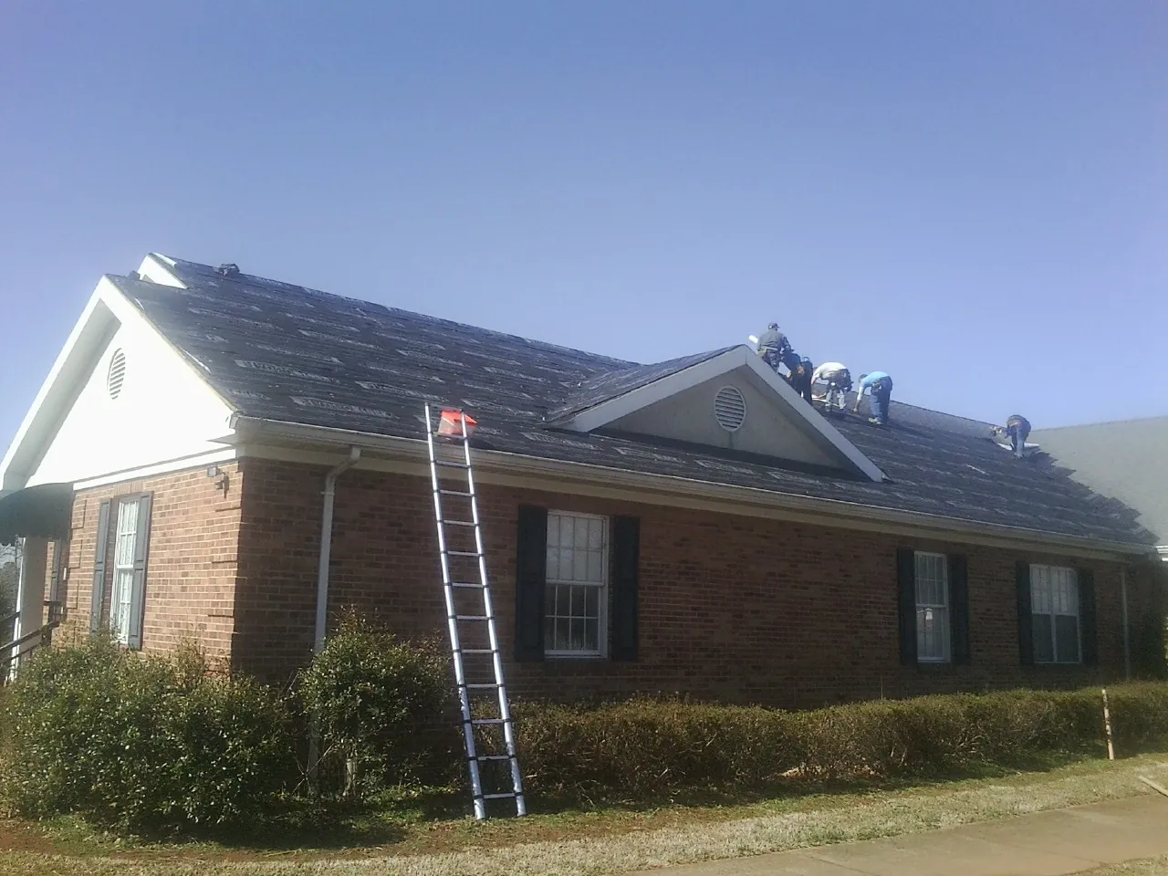 Crew tearing shingles off a brick ranch roof with a ladder set — Emergency Roof Repair work by our crew in Bertha