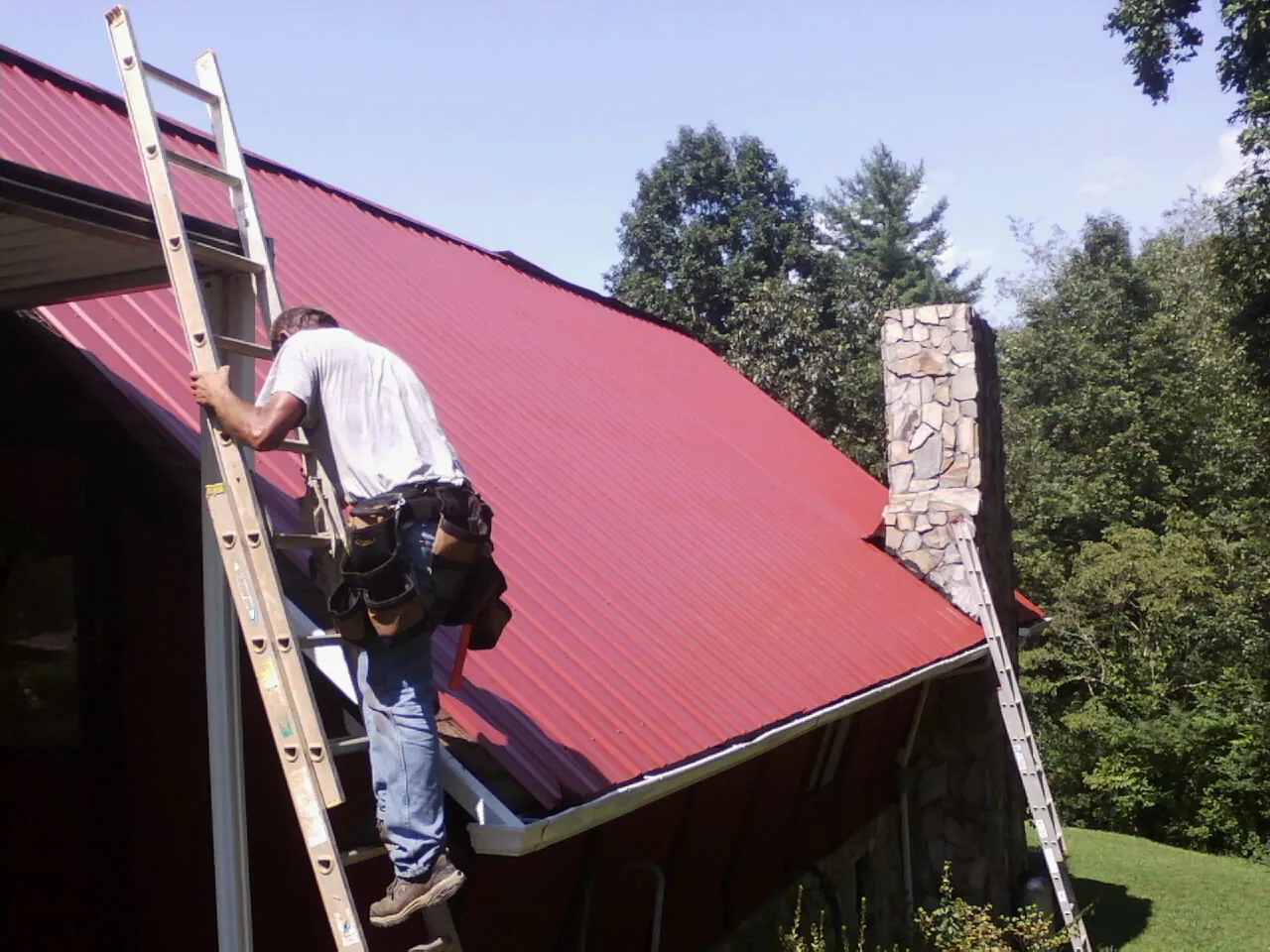 Roofer on a ladder beside a red metal roof and stone chimney — Soffit & Fascia Repair by Roofing Brothers in Bertha